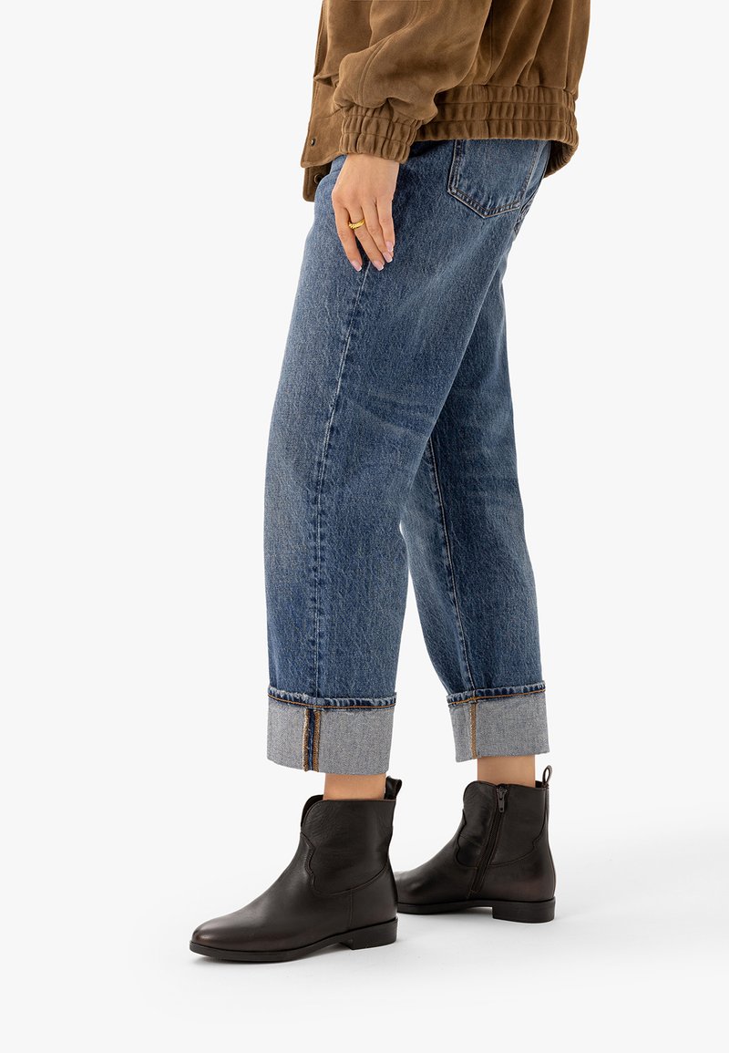 Personne portant un jean bleu retroussé, une veste marron, des bagues dorées et des bottines en cuir noir, debout sur un fond blanc.
