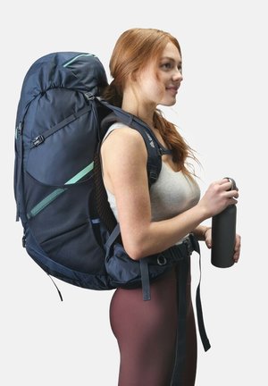 Navy blue backpack with green accents, padded straps, and mesh fabric. Features adjustable buckles and is being held by a person with a water bottle.