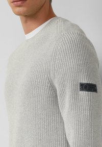 Hellgrauer, strukturiert gestrickter Pullover mit Rundhalsausschnitt, versehen mit einem schwarzen Logo-Patch auf dem linken Ärmel und einem weißen Innenkragen.
