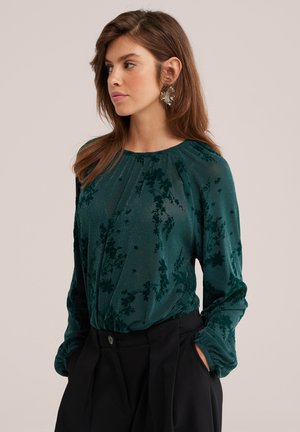 Femme aux cheveux bruns portant un chemisier vert à fleurs à manches longues et un pantalon noir, debout les mains dans les poches, regardant vers la gauche.