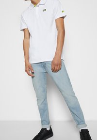 Biała koszulka polo z zielonym akcentem na kołnierzyku, z logo na klatce piersiowej. W połączeniu z jasnoniebieskimi dżinsami i czarnymi trampkami.
