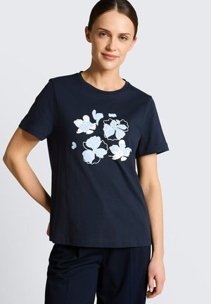 Kobieta w granatowej koszulce z jasnoniebieskimi i białymi kwiatowymi wzorami, stojąca z jedną ręką na biodrze na jasnym tle.