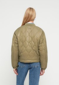 Καπιτονέ bomber μπουφάν σε ελιά πράσινο με ριμπ γιακά. Διακρίνονται στρογγυλά σχέδια και εφαρμοστά μανσέτα, συνδυασμένα με ψηλόμεσα μπλε τζιν.