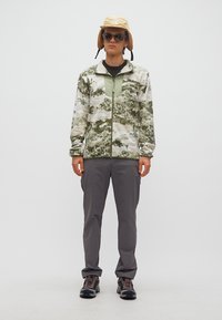 Άτομο με φλις μπουφάν παραλλαγής, γκρι παντελόνι, παπούτσια πεζοπορίας, μπεζ καπέλο τύπου κάλπη με πλαϊνά καλύμματα αυτιών και μαύρα γυαλιά ηλίου, στέκεται μπροστά σε λευκό φόντο.