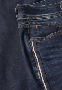 Granatowe dżinsy z klasycznym pięciokomorowym wzorem, widoczne szwy w kontrastującym złotym kolorze oraz białe paseczki podszewki kieszeni.