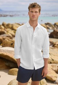 White linen button-up shirt with rolled sleeves and a chest pocket, paired with navy swim shorts, against a rocky beach background.