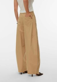 Personne portant un pantalon beige à jambes larges et des talons pointus marron, avec une main dans la poche arrière, debout devant un fond uni.