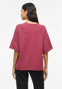 Sieviete ar gariem tumšiem matiem, ģērbusies vaļīgā tumši rozā īsroku džemperī un melnās biksēs, redzama no muguras.