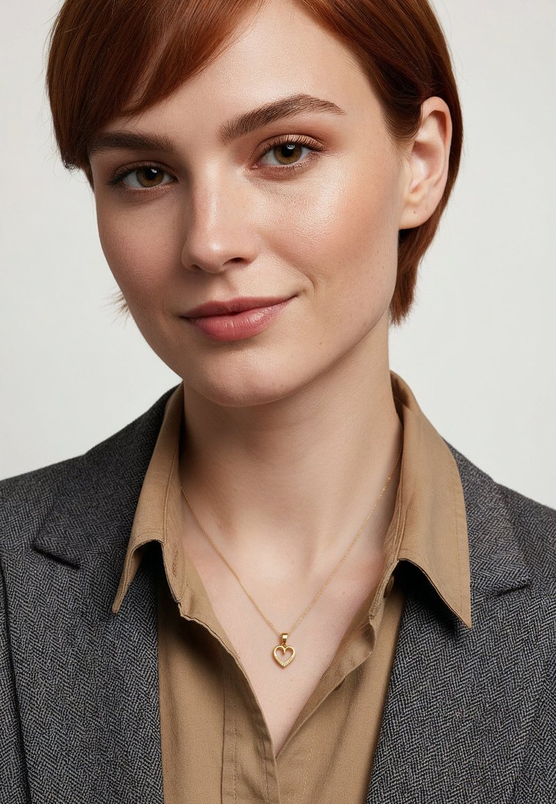 Goldenes Herzanhänger-Halskette an einer zarten Kette, getragen über einem beigen Hemd mit Kragen und einem grauen Fischgrät-Blazer. Dezenter Glanz und filigranes Design.