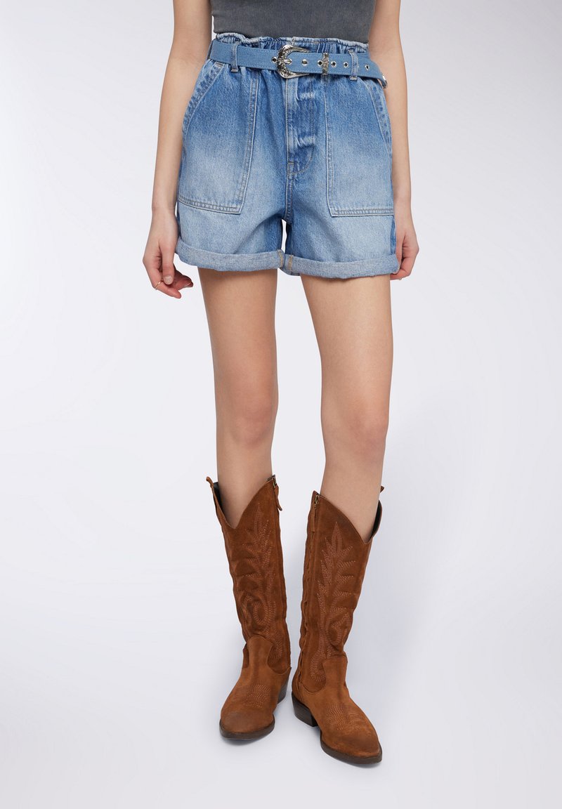 Shorts di denim con vita alta, orli arrotolati e due tasche frontali. Abbinati a stivali cowboy alti in suede marrone con dettagli di cucitura.