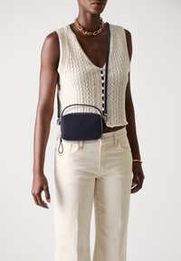 Geantă de umăr din piele de marină cu logo embosat, formă dreptunghiulară și componente din metal argintiu; purtată cu un top fără mâneci din tricot și pantaloni crem.