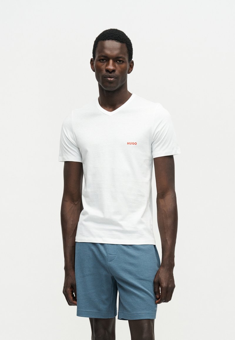 Άνδρας στέκεται φορώντας λευκό T-shirt με V-λαιμόκοψη με το κείμενο «HUGO» και μπλε υφασμένα σορτς σε απλό λευκό φόντο.