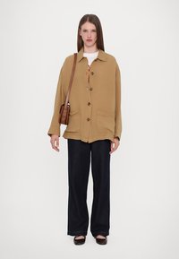 Khaki oversized jacket with large pockets, button closure, and long sleeves, paired with dark wide-leg pants and a brown shoulder bag.