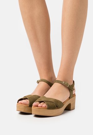 Sandali platform in suede verde oliva con tacco in legno spesso, punta aperta e cinturino alla caviglia con dettaglio in metallo.