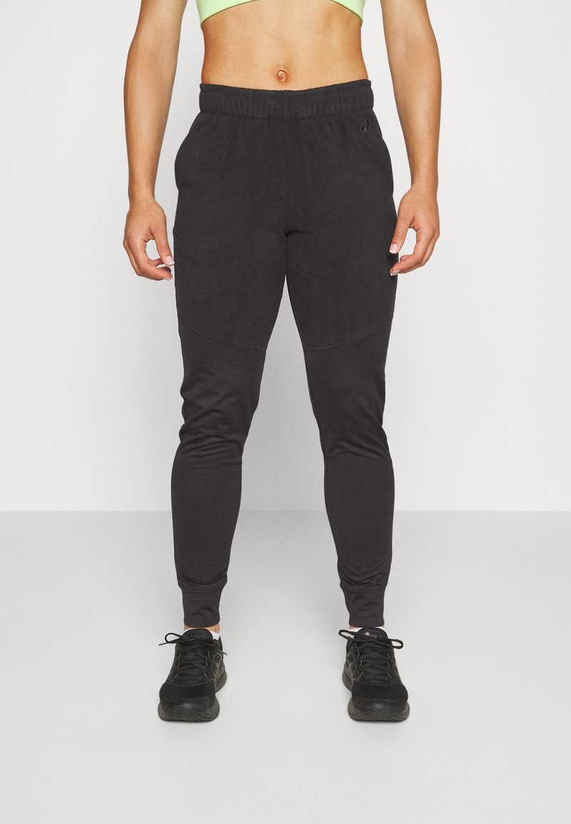 Zwarte fleece joggingbroek met een elastische tailleband, taps toelopende enkels en zijzakken, gecombineerd met zwarte sportschoenen op een effen achtergrond.