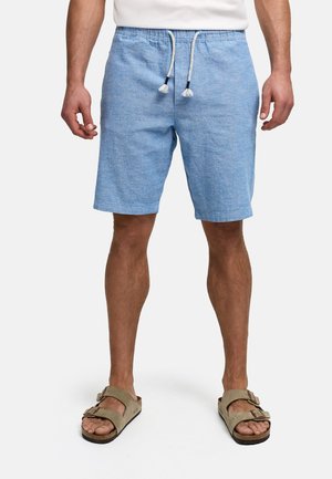 Mand iført lyseblå snøreshorts og beige sandaler med to stropper, stående foran en ensfarvet hvid baggrund.