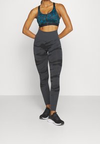 Reggiseno sportivo con stampa leopardata turchese, leggings camouflage grigi a vita alta e scarpe da ginnastica nere. La texture è liscia e il design è aderente.