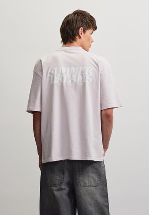Jeune homme portant un t-shirt oversize rose clair avec le texte "SUMMER DREAMS" dans le dos et un jean sombre et ample, debout de profil.