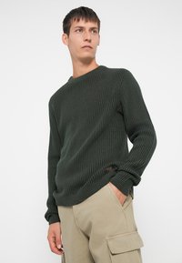 Junger Mann, der einen dunkelgrünen gerippten Pullover und beige Cargo-Hosen trägt, mit einer Hand in der Tasche, der zur Seite blickt, vor einem weißen Hintergrund.