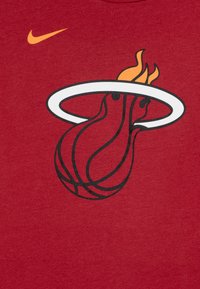 Sarkana kokvilnas T-krekls ar melnu un oranžu Miami Heat logo, kas attēlo liesmojošu basketbola bumbu. Nike swoosh logo augšpusē oranžā krāsā.