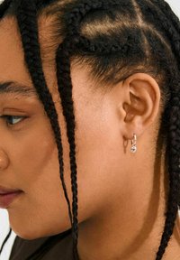 Silver hoops med en liten hängande charm, bärs i en flätad frisyr, som visar textur och subtil glans mot huden.