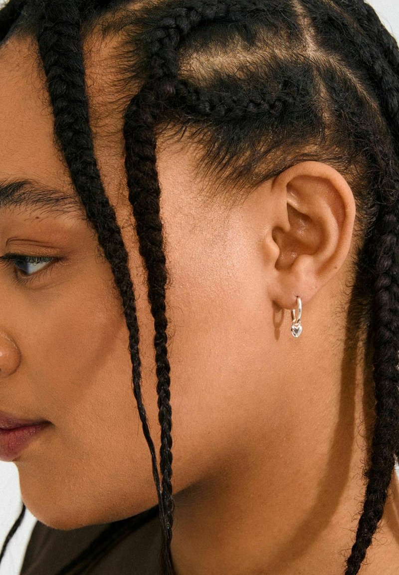 Silver hoops med en liten hängande charm, bärs i en flätad frisyr, som visar textur och subtil glans mot huden.