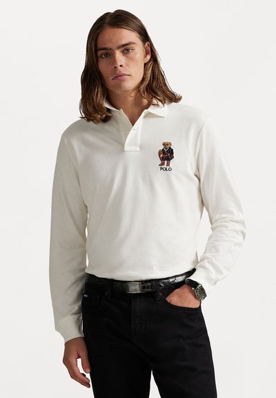 Polo de manga larga en crema con cuello, que presenta un oso bordado y el logo "POLO" en el lado izquierdo del pecho, combinado con pantalones oscuros.