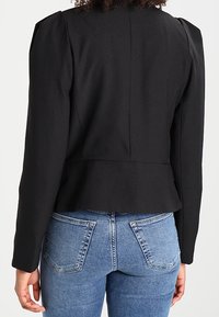 Blazer noir sur mesure avec épaules bouffantes, taille cintrée et design péplum, associé à un jean bleu clair.