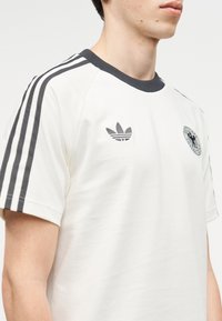 Fiatal férfi fehér Adidas pólóban, fekete három csíkkal az ujjakon és logóval a mellkason, egyszínű háttér előtt.