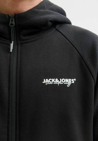 Zwarte zip-up hoodie met een gestructureerde kap en schouderaccenten. Beschikt over een geborduurd logo, ribbeldetails en een ritssluiting aan de voorkant.