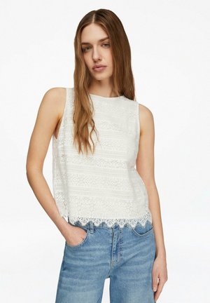 Donna con capelli lunghi castano chiaro, indossa una canotta bianca di pizzo e jeans blu, in piedi con una mano in tasca.