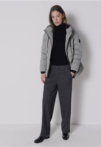 Grå vadderad jacka med huva över en svart polotröja, tillsammans med mörkgrå byxor och svarta skor, allt mot en neutral bakgrund.