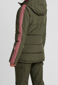 En person som bär en mörkgrön puffjacka med rosa ränder står vänd bort. Jackan har en huva och dragkedjefickor.