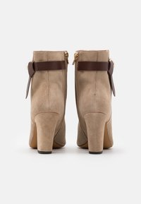 Un par de botines de ante color topo con tacones de bloque, detalle de correa de cuero marrón y cremallera dorada en la parte trasera, mostrados desde la vista trasera.