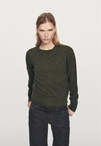Olivengrøn, strikket sweater med V-hals, tætsiddende design og lange ærmer. Båret over et mørkegrønt lag, kombineret med mørke denim jeans.
