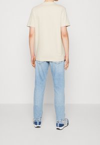 Camiseta de algodón beige, de manga corta, con cuello redondo, combinada con jeans de mezclilla azul claro y zapatillas en azul, gris y blanco.
