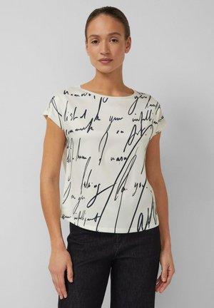 Kobieta ubrana w biały krótki rękaw z czarnym abstrakcyjnym ręcznie pisanym napisem oraz czarne spodnie na jednolitym tle.