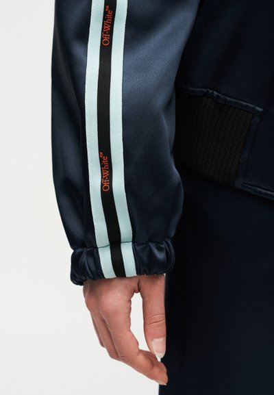 Χέρι δίπλα σε μανίκι ναυτικού μπλε ζακέτας με λεπτομέρειες σε ανοιχτό μπλε και μαύρες ρίγες και μικρά πορτοκαλί λογότυπα "Off-White" στο μανίκι.