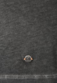 Tmavošedá tkanina s jemnou strukturou, s kruhovou kovovou nášivkou s reliéfním logem, připevněná oranžovou nití.