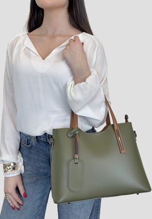 Donna in camicia bianca e jeans blu che tiene una grande borsa verde oliva con manici color cuoio e cartellino attaccato.