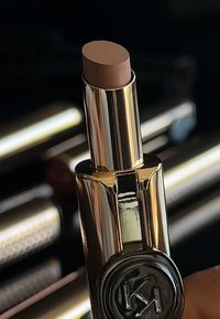 Pomadka w złotej metalowej obudowie z okrągłym czarnym emblematem. Kulka ma płaską górę w beżowo-brązowym odcieniu. Tło z rozmytymi srebrnymi tubkami.