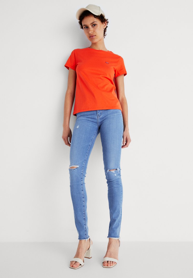 Helles orange T-Shirt, kurze Ärmel, Rundhals; kombiniert mit hellblauen Skinny Jeans mit Rissen. Das Model trägt beige Absatzsandalen.