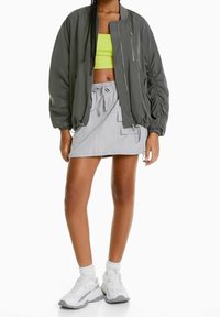 Šedá bomber bunda se zapínáním na zip, spárovaná s neonově zeleným crop topem a světle šedou sukní s kapsami. Bílá sneakers dokončuje vzhled.