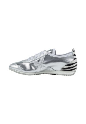 Zapatilla deportiva plateada de perfil bajo con cordones y suela blancas, y detalle de talón a rayas negras y blancas, mostrada orientada hacia la izquierda sobre fondo blanco.