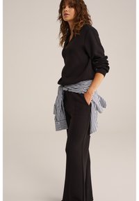 Femeie stând lateral, purtând un top negru cu mâneci lungi, pantaloni negri și o cămașă cu dungi alb-negru legată în jurul taliei.