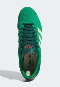 Green suede sneaker with cream stripes, textured tongue, red logo detail, and dark green accents. Flat laces and padded collar design.