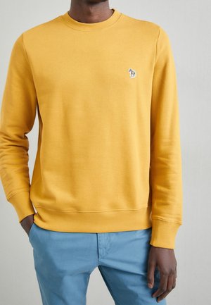 Sudadera amarilla con cuello redondo, puños y dobladillo acanalados; cuenta con un pequeño logo bordado en el pecho. Combinada con unos pantalones azul claro.