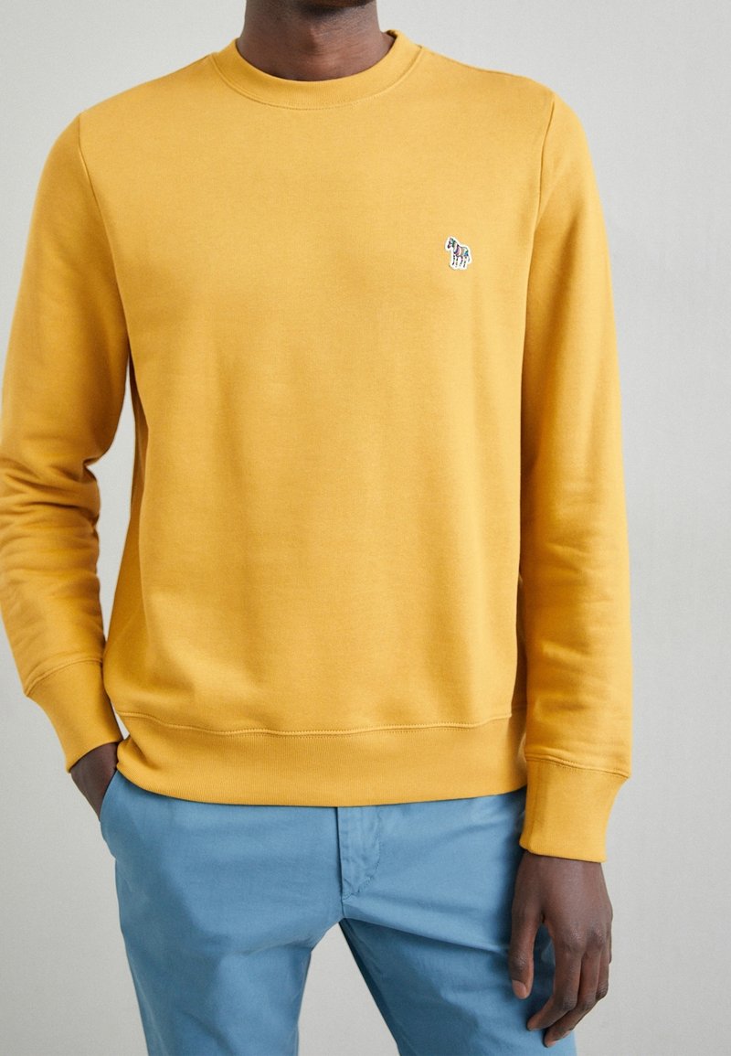 Sudadera amarilla con cuello redondo, puños y dobladillo acanalados; cuenta con un pequeño logo bordado en el pecho. Combinada con unos pantalones azul claro.