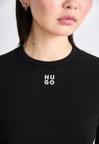 Crna pamučna majica s okruglim izrezom i bijelim izvezenim "HU" i "GO" na prsima. Glatka tekstura, kroj koji se prilagođava tijelu.
