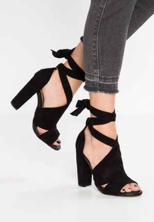 Beine in grauen, kurzen Jeans, die schwarze Wildleder-High Heels mit dicken Blockabsätzen und Wickelriemen um die Knöchel tragen, die zu Schleifen gebunden sind.